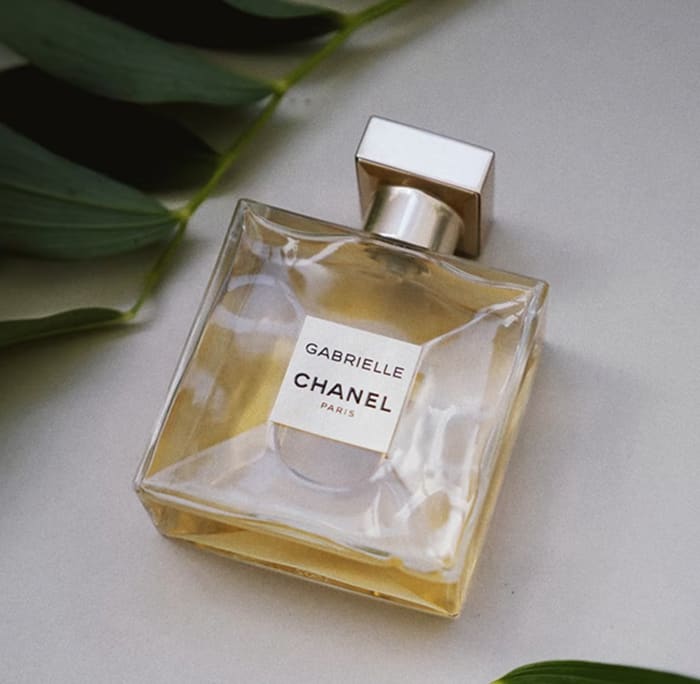 Gabrielle Chanel Essence Eau de Parfum - elegant glass bottle with gold liquid surrounded by green tropical leaves